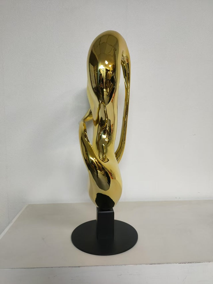 Gold abstract sculpture on a black stand against a white background