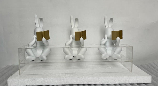 Three white rabbit sculptures with gold books on a gray surface and light gray background