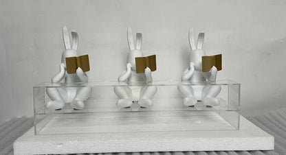 Three white rabbit sculptures with gold books on a gray surface and light gray background