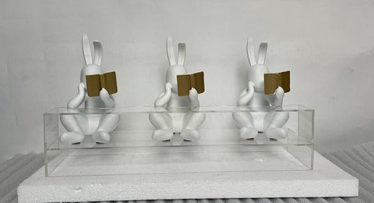 Three white rabbit sculptures with gold books on a gray surface and light gray background