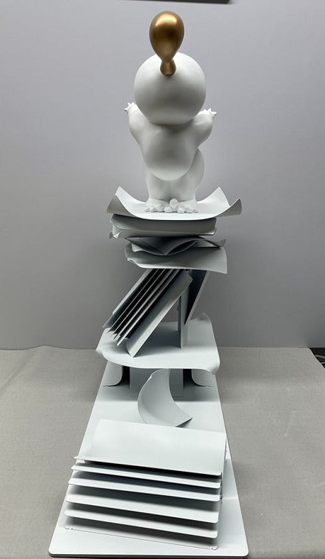 White ceramic sculpture with a gold ball on top, placed on a white surface.
