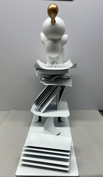 White ceramic sculpture with a gold ball on top, placed on a white surface.