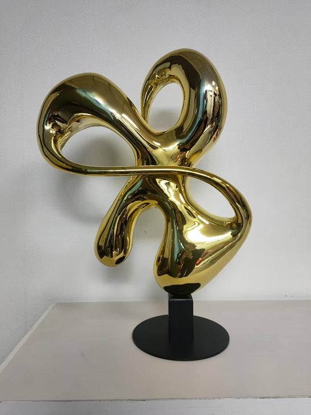 Gold abstract sculpture on a black stand against a white background