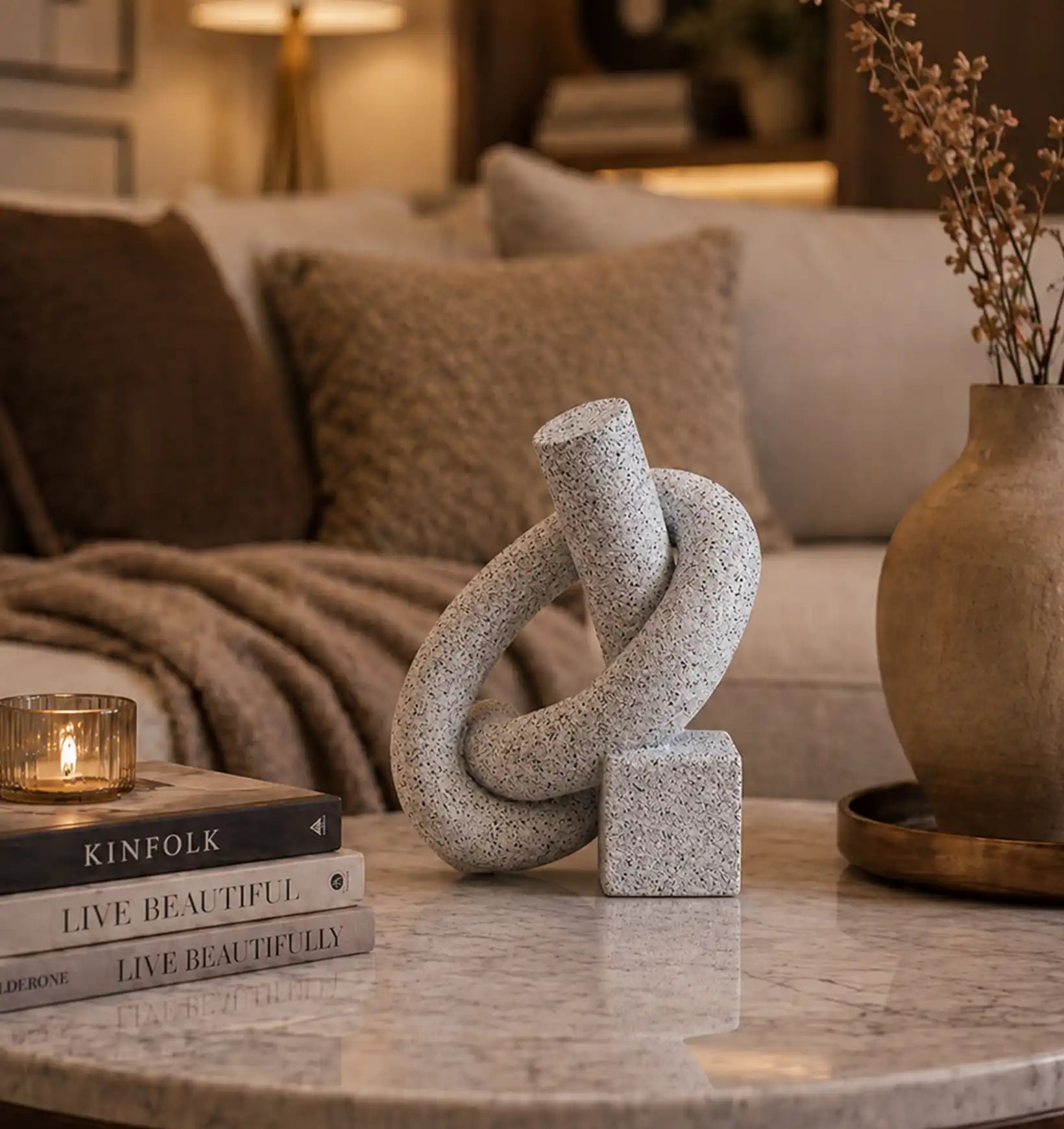 Grey knot sculpture on a marble table with books, a candle, and a vase with dried flowers